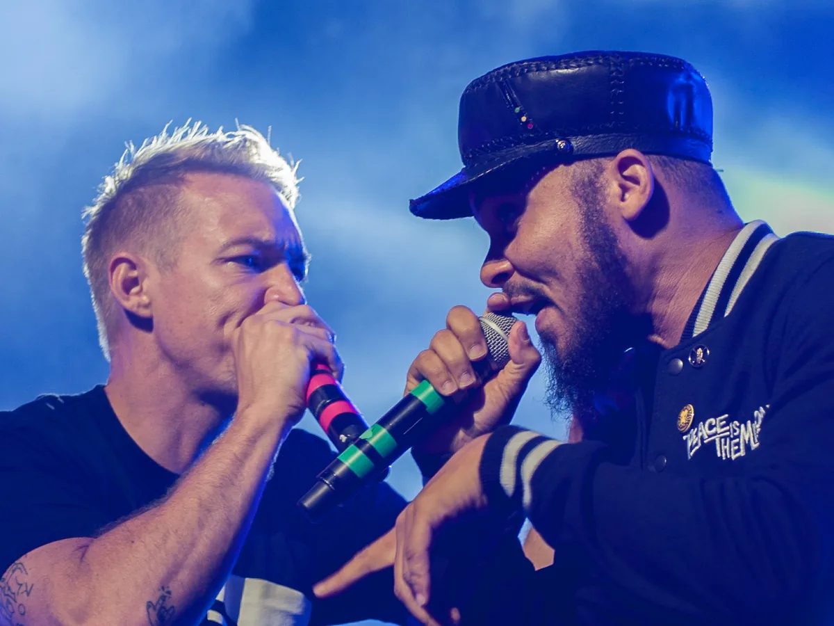 Diplo, bleached hair and a dark T-shirt, and Walshy Fire, in a cap and varsity jacket, shouting into microphones face to face under blue stage light