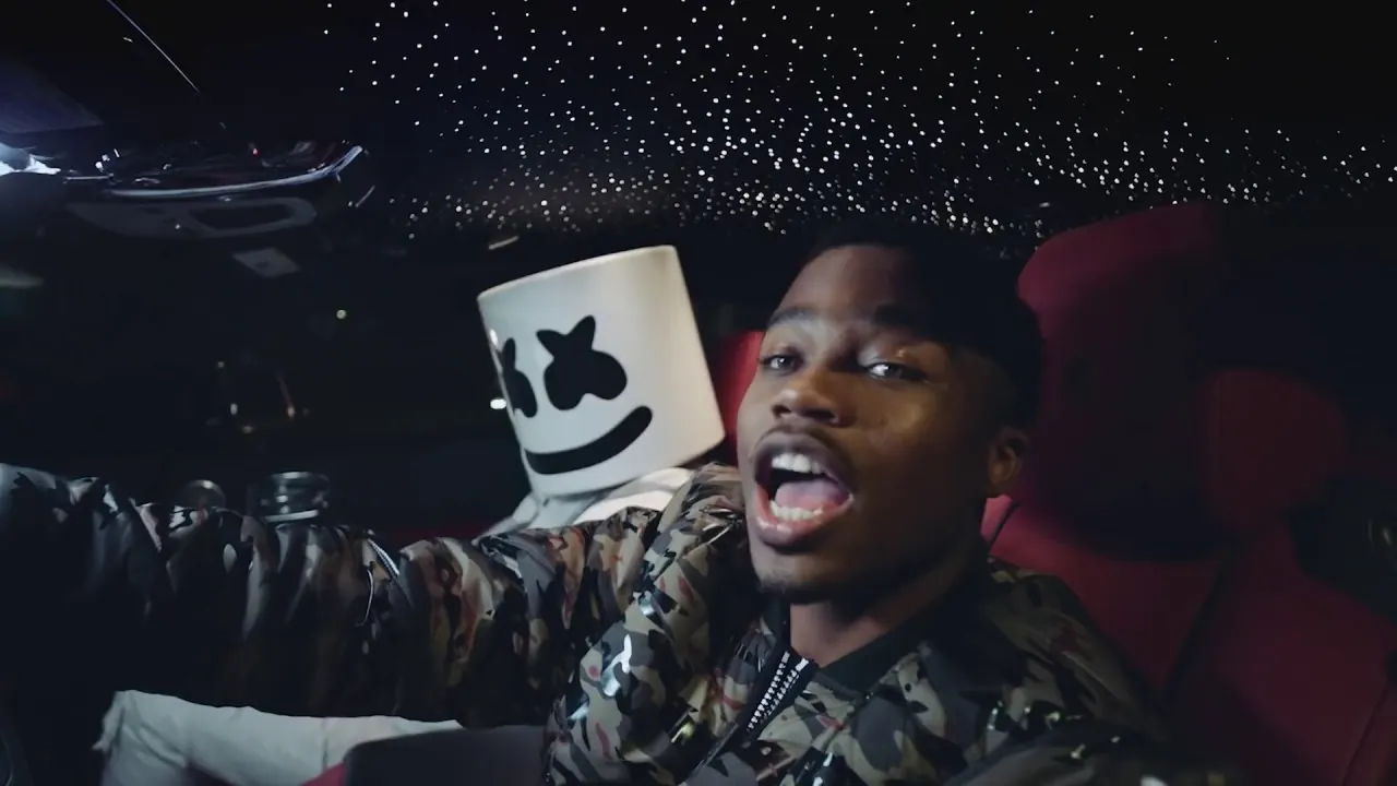 Roddy Ricch rapping in the back seat of a car with a starlit headliner, Marshmello in his white helmet beside him