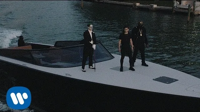 Three men standing on the deck of a speedboat on a canal at dusk, one in Joker makeup and a white suit with a cane