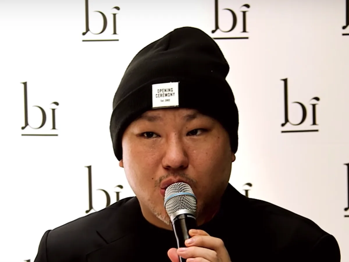 Brave Brothers in a black beanie and jacket, speaking into a microphone in front of a logo backdrop