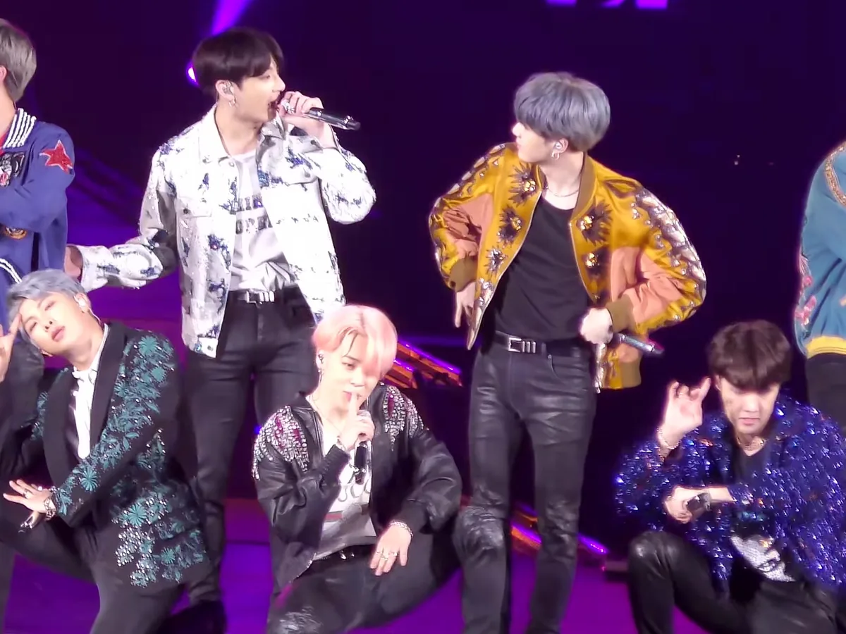 The seven members of BTS in sequined and printed jackets, several crouching, performing on a purple-lit arena stage