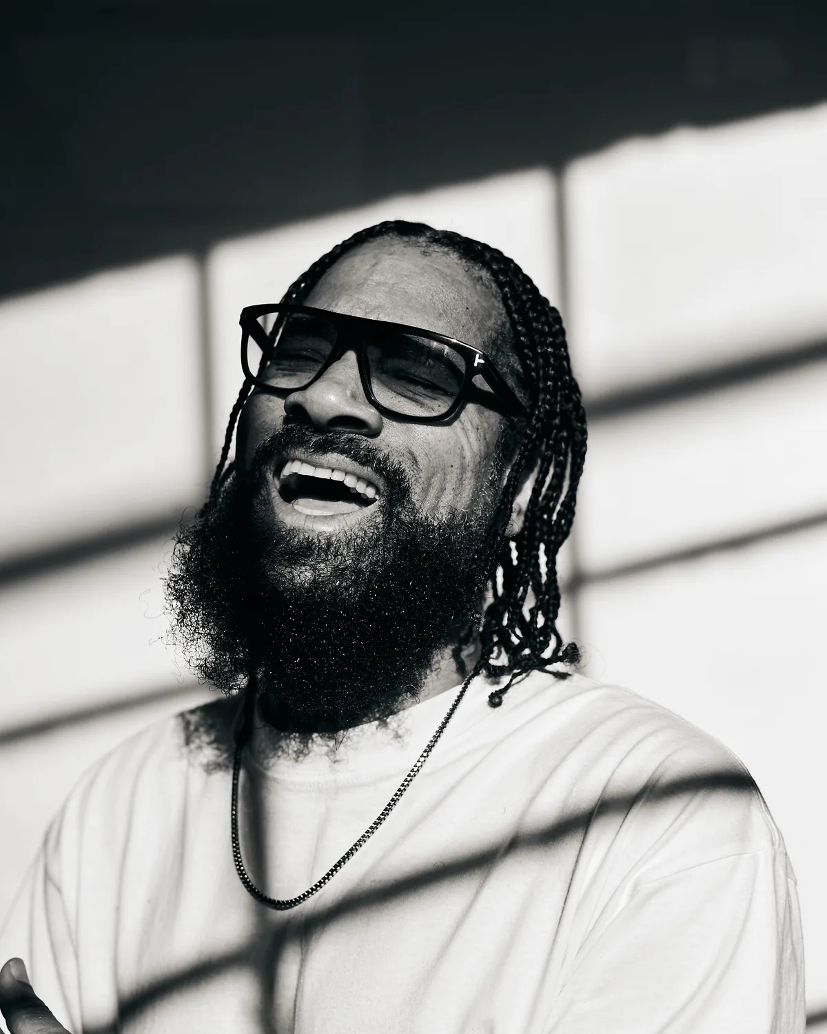 Black-and-white portrait of Dem Jointz in glasses and a white sweatshirt, laughing, with braided hair and a full beard