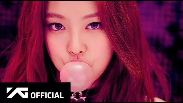 Close-up of Jennie with long red-tinted hair blowing a pink bubble of gum, beside the YG Official logo