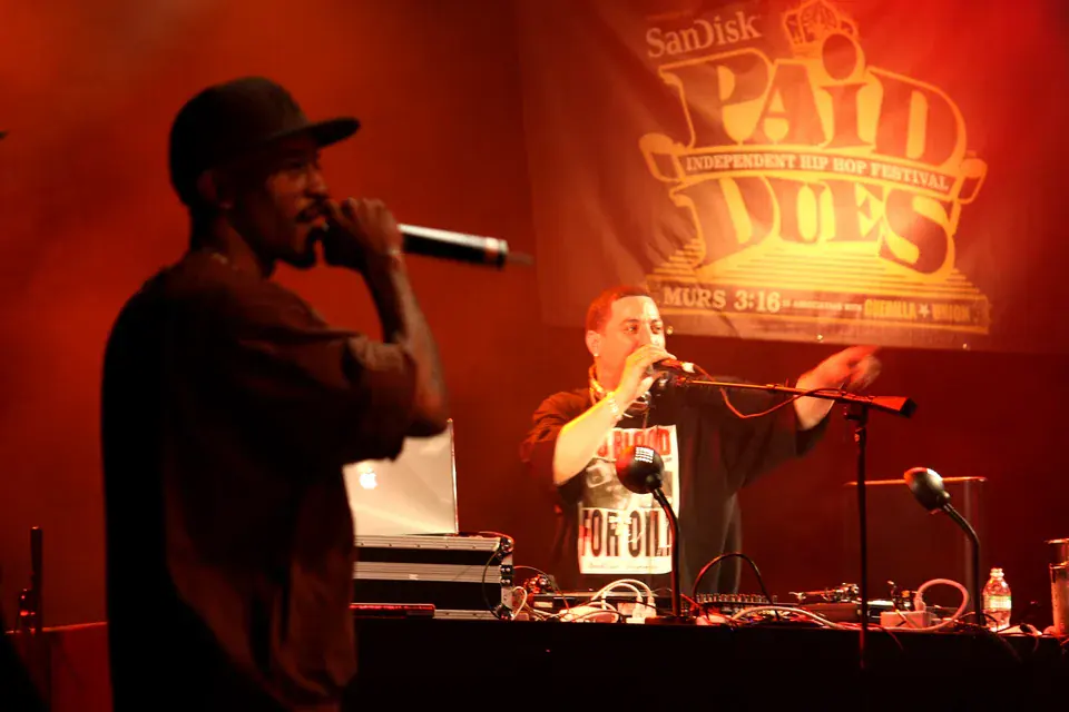 Rakim at the microphone in red stage light while Kid Capri works the turntables under a Paid Dues banner