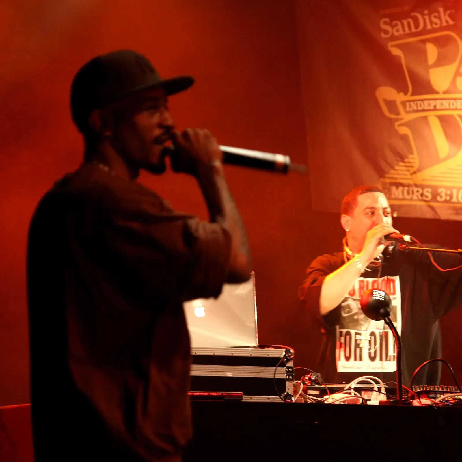 Rakim performs into a microphone in red stage light while Kid Capri works the turntables behind him under a Paid Dues festival banner