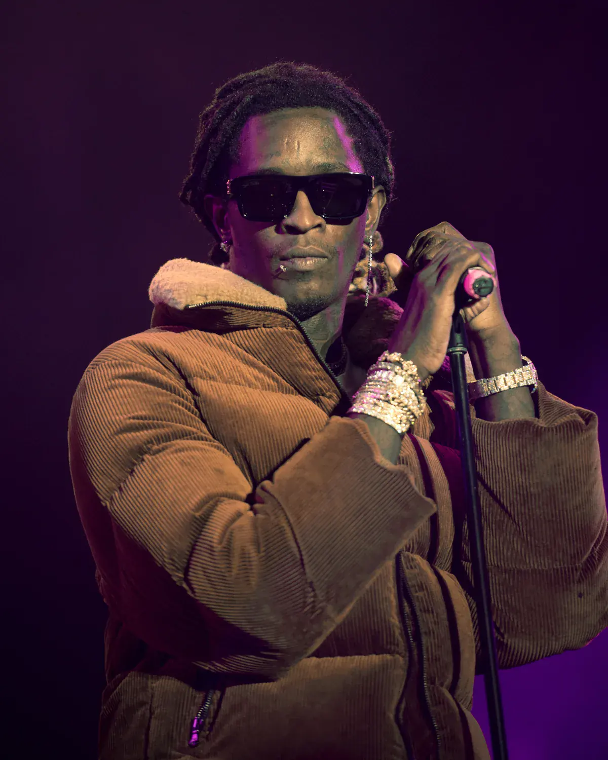 Young Thug in sunglasses and a brown jacket holds a microphone to his mouth under purple stage light