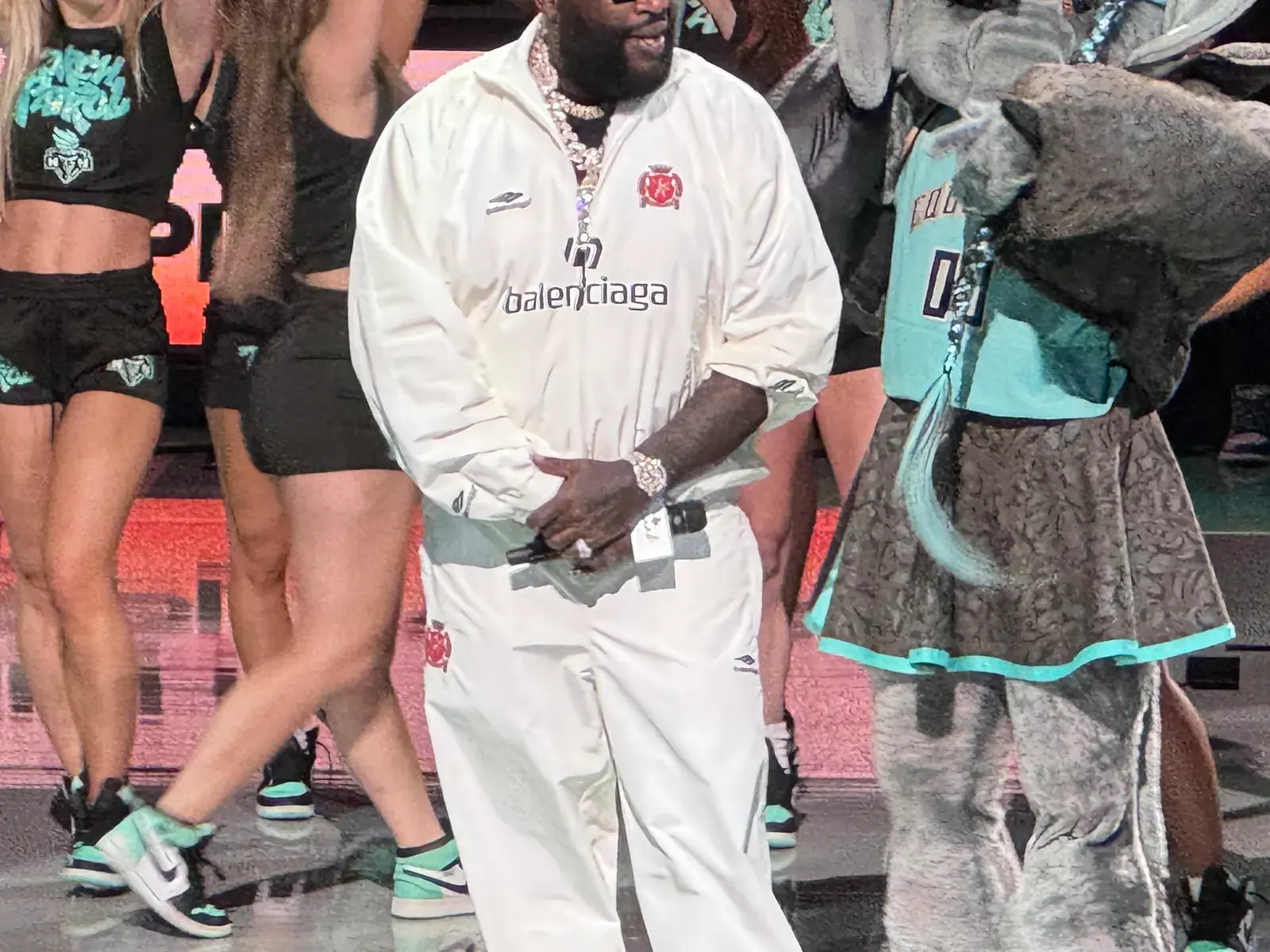 Rick Ross in white performing on a basketball court with dancers waving towels and a team mascot