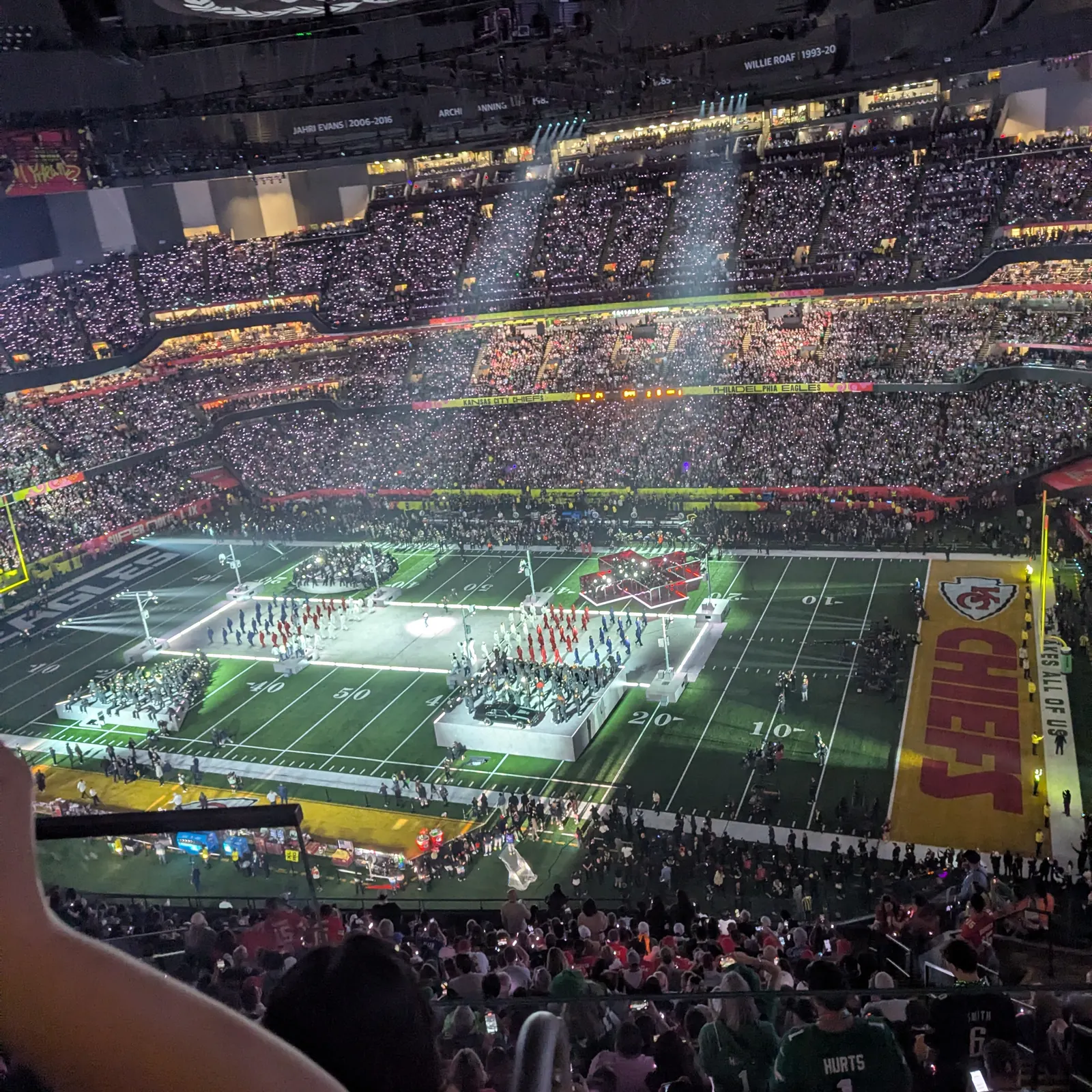 A packed stadium seen from the upper stands, the field lit for a halftime show with crowds of performers on it