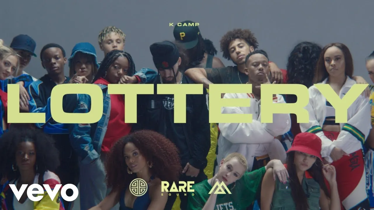 A crowd of young dancers in streetwear posing against a grey wall behind the word LOTTERY in large yellow letters, with K Camp’s name above it