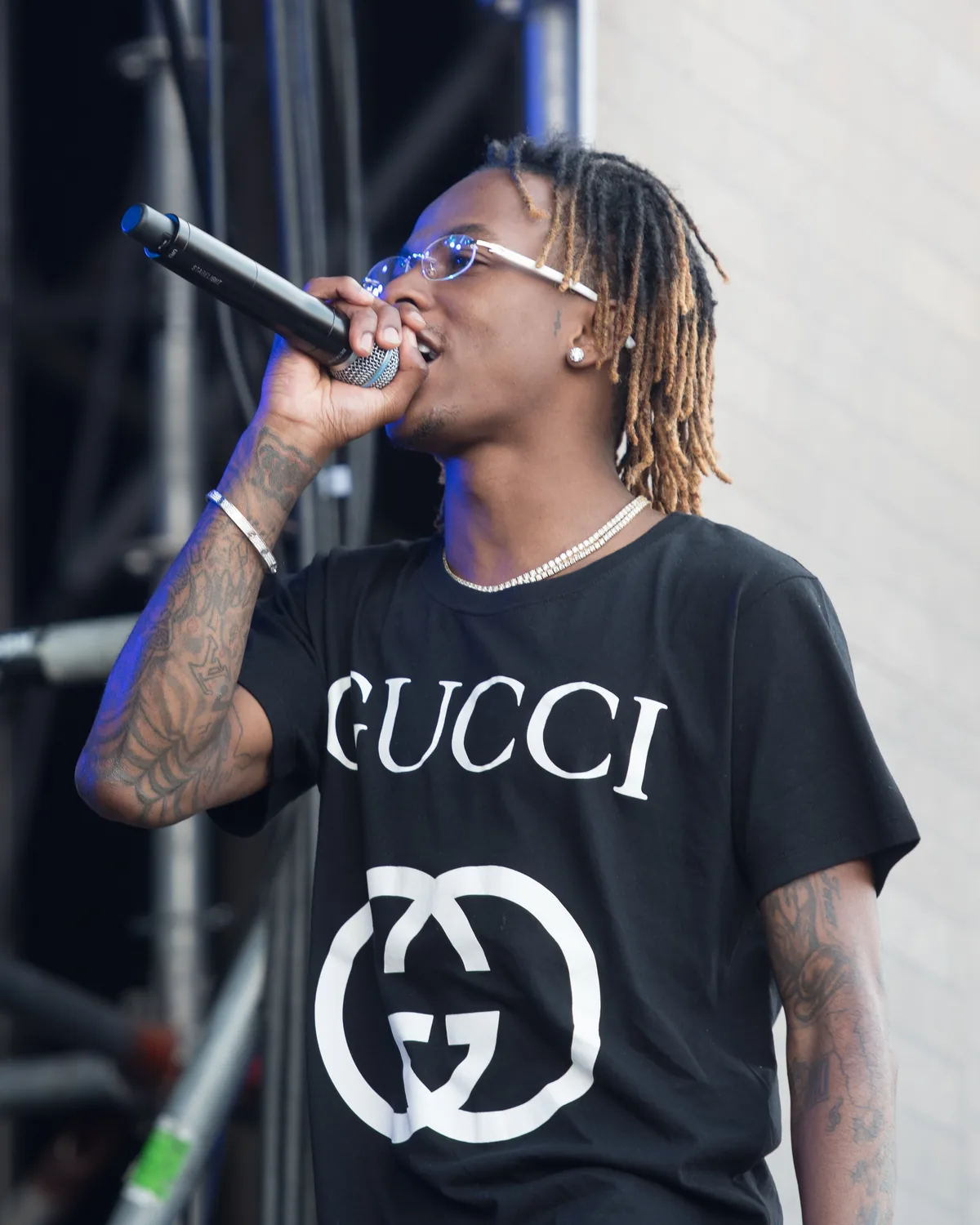 Rich The Kid in sunglasses and a black Gucci T-shirt, rapping into a handheld microphone on an outdoor stage