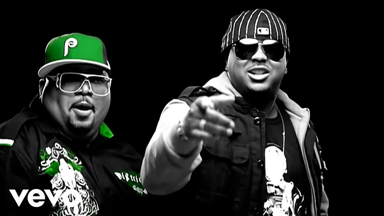 Two men in sunglasses against black in a black-and-white frame with green accents, one in a green cap, the other in a striped cap pointing at the camera