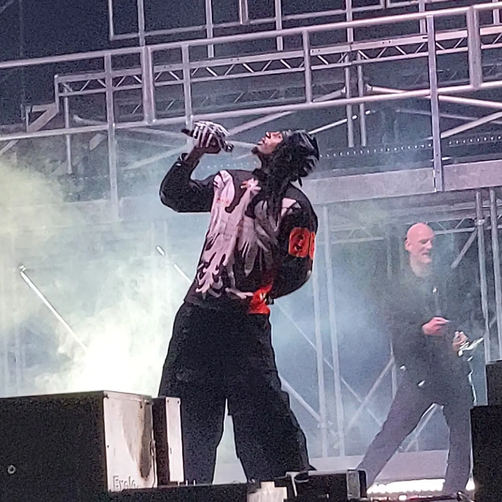 Playboi Carti in a black hood and a jersey with a white eagle, head thrown back and microphone raised, under stage scaffolding and smoke