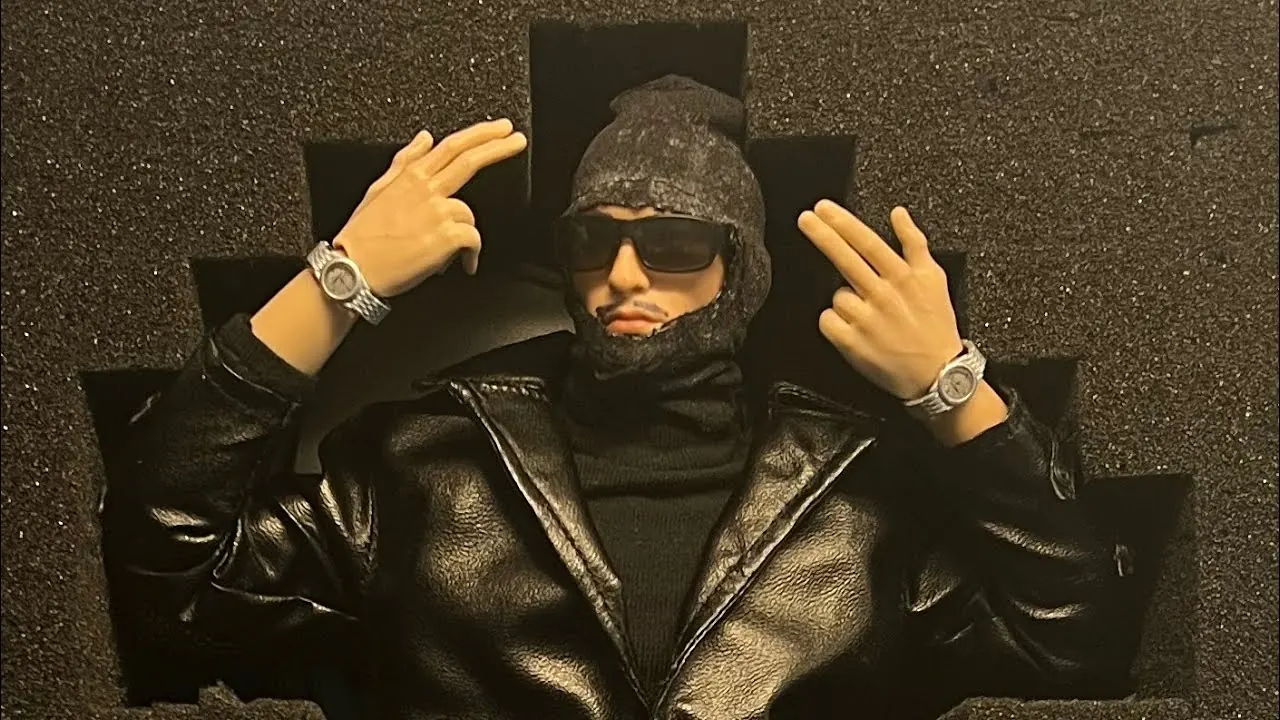 An action figure in a black balaclava, dark sunglasses and a leather coat, posed in black foam packaging