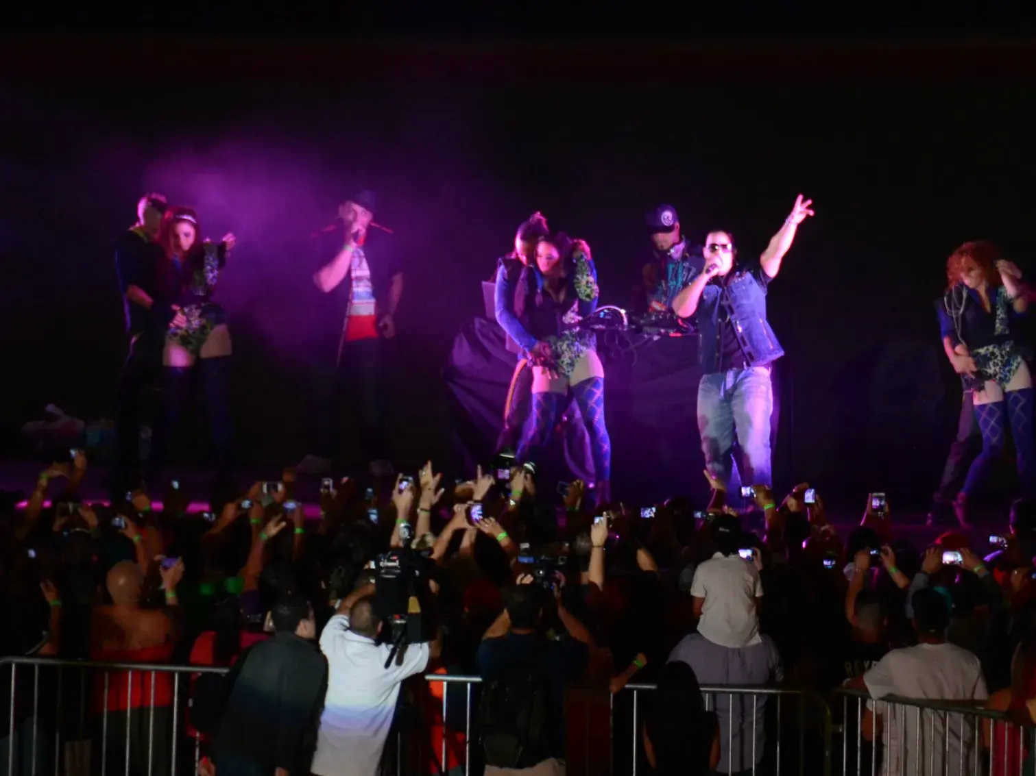Daddy Yankee and dancers on a stage lit purple, arms raised, above a crowd holding up phones