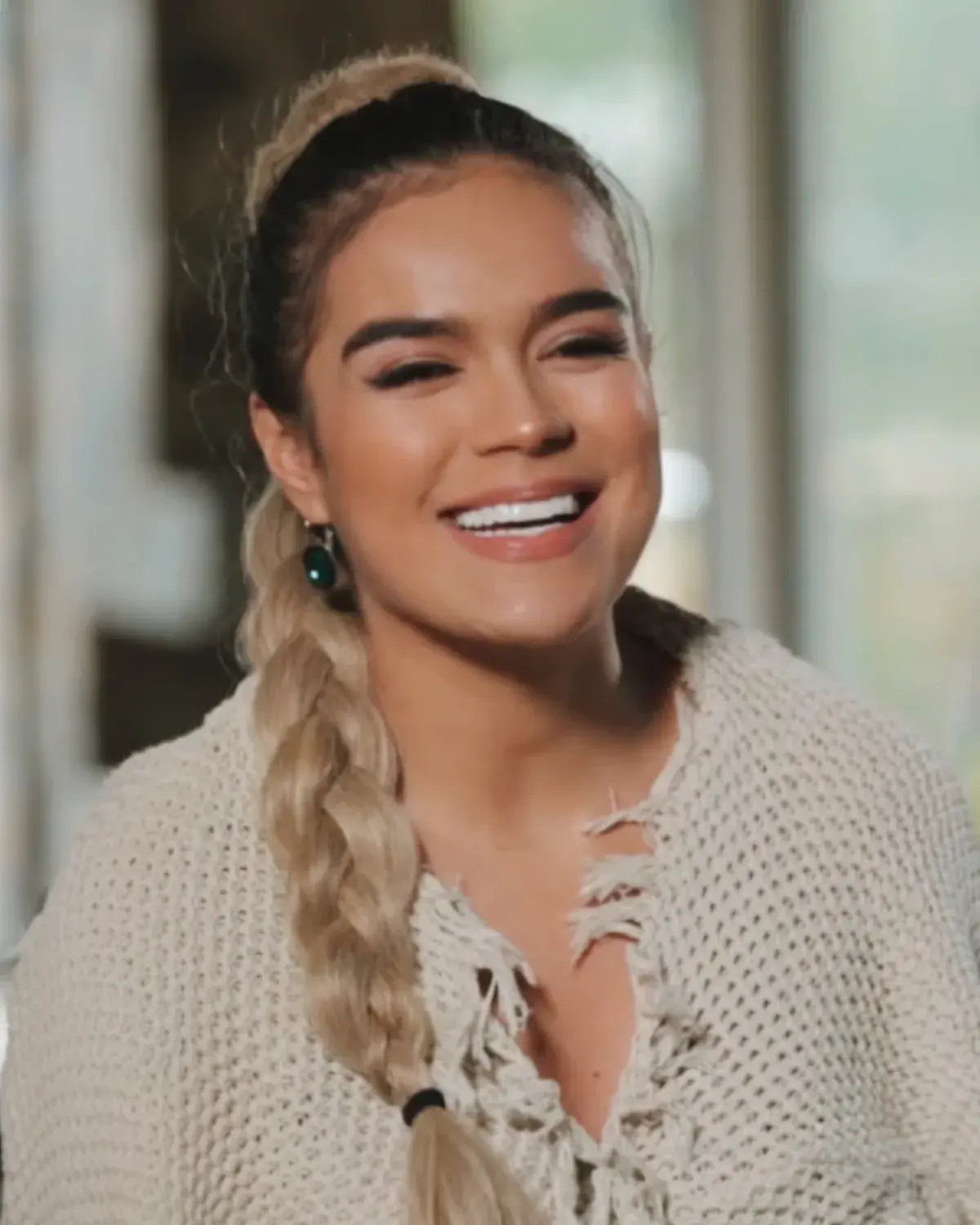 Karol G smiling, with a long braid over her shoulder and a cream knit sweater, seated for an interview