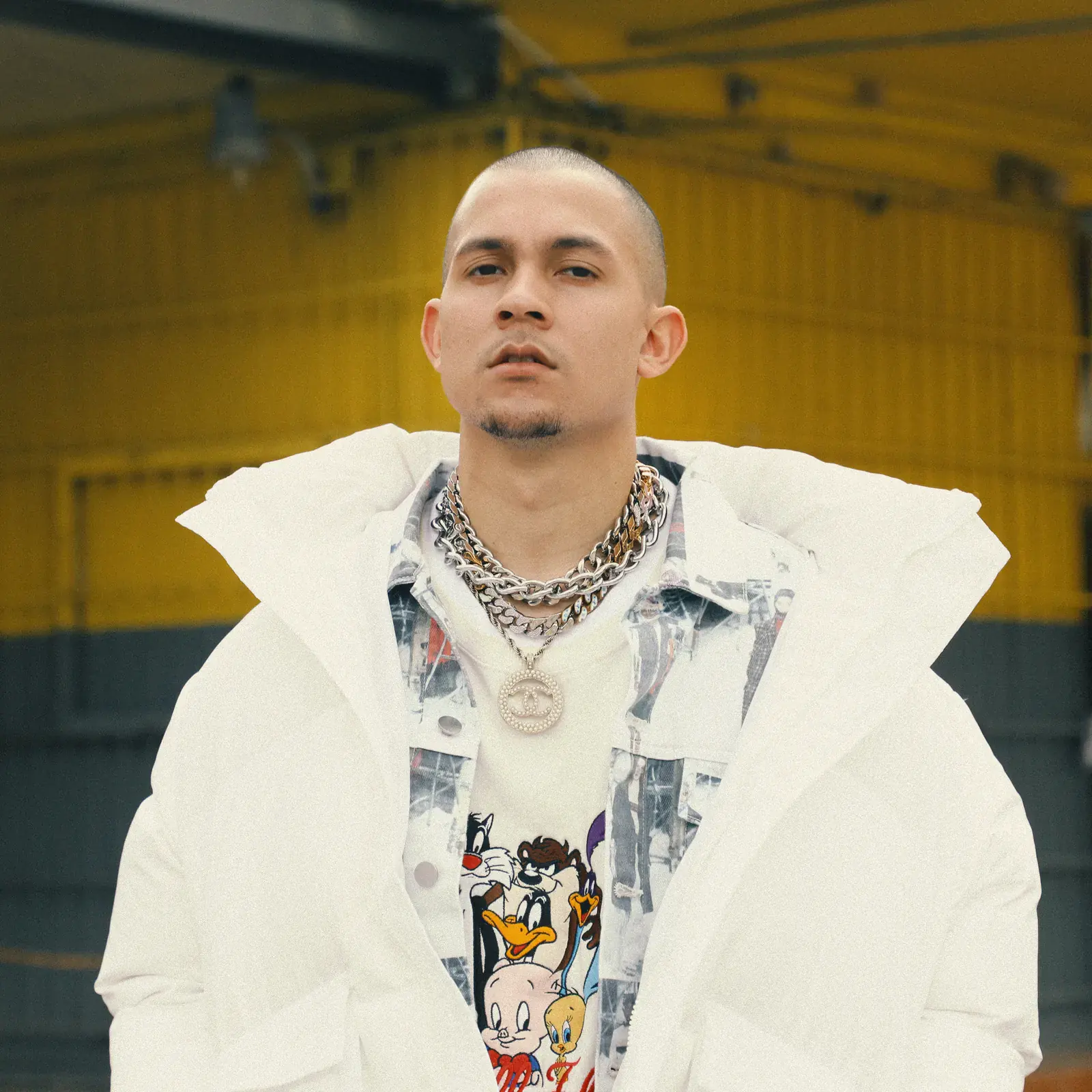 Tainy in a white puffer coat and chains over a T-shirt printed with Looney Tunes characters, in front of a yellow wall