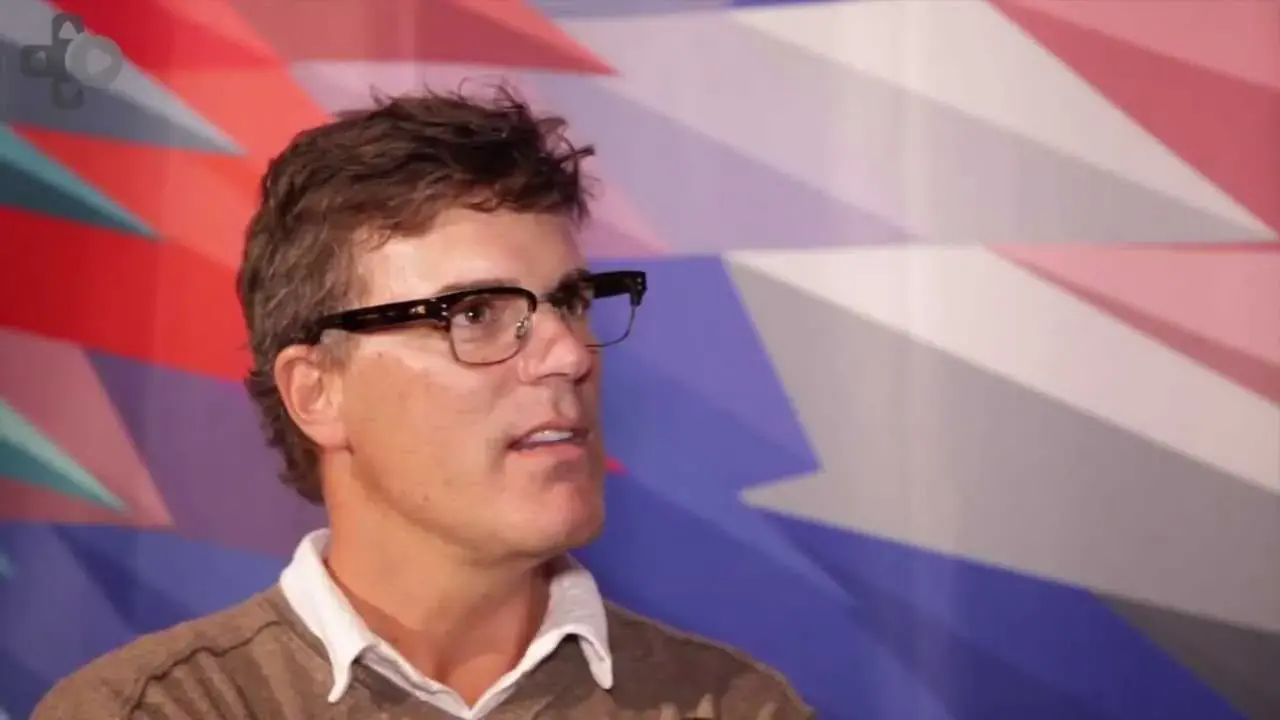 Andrew Anthony in glasses and a sweater, speaking in front of a colorful geometric wall
