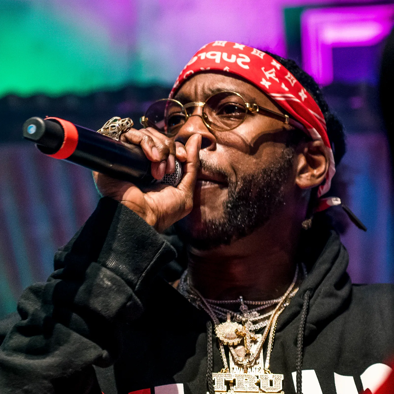 2 Chainz in a red bandana and round glasses raps into a microphone, one finger raised to his lips